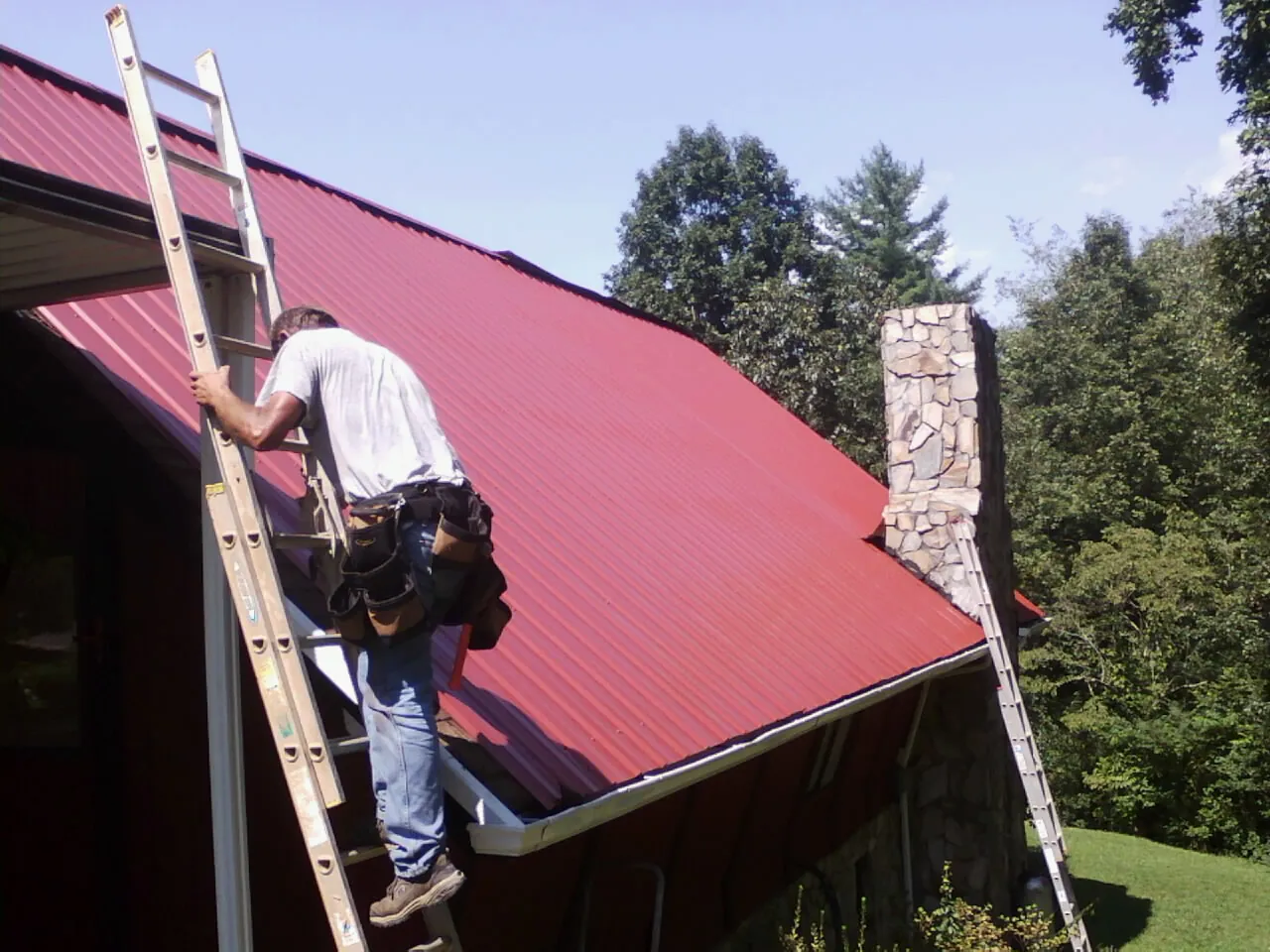 Roofer on a ladder beside a red metal roof and stone chimney — Soffit & Fascia Repair by Roofing Brothers in Seneca Falls